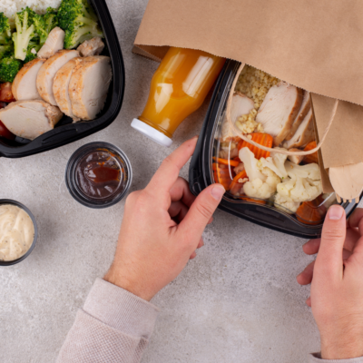 Osoba rozpakowująca jedzenie na wynos z papierowej torby, zawierające kurczaka, warzywa, bulgur oraz butelkę soku.