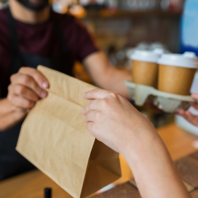 Osoba odbierająca papierową torbę z jedzeniem oraz napoje na wynos w kubkach od pracownika restauracji.