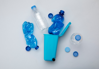 Różne plastikowe butelki (PET) i nakrętki wokół małego niebieskiego kosza na śmieci, symbolizujące system kaucyjny i recykling opakowań.